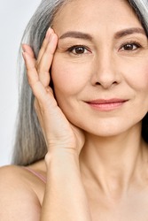 Vertical portrait of middle aged Asian woman's face with perfect skin. Older mature lady touching pampering face with hand. Advertising of cosmetology salon rejuvenating spa procedures skincare.