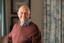 Portrait of happy retired senior man standing at home near window. Satisfied old man looking at camera and smiling while standing near the window. Positive and confident elderly enjoy his retirement.