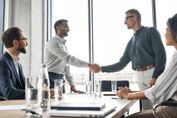 Two happy diverse professional business men executive leaders shaking hands after successful financial deal at group board office meeting. Trust agreement company trade partnership handshake concept.