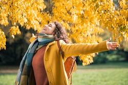 Beautiful young woman relaxing at park during autumn season with closed eyes. Happy free natural girl breathing deeply in park with foliage in background. Pretty woman expressing freedom outdoor.