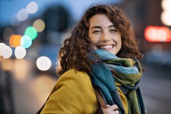 Portrait of happy woman on city street looking at camera. Beautiful girl standing outdoors in winter evening with a toothy smile. Cheerful university student wearing coat with woolen scarf at dusk.