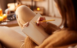 halloween, holidays and leisure concept - close up of young woman with pencil writing to diary and resting her feet on table at cozy home