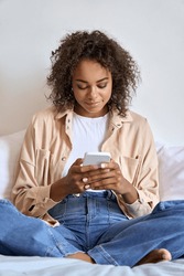 Young happy African American woman using smart phone sitting on bed. Smiling girl holding cellphone online mobile apps, texting in chats, searching web technology relaxing in bedroom.