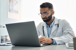 Indian male doctor medic expert wearing white coat using laptop computer at work in hospital, checking patients electronic files medical forms, having online virtual telehealth consultation.