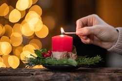 Close up of woman hand lighting red candle with a match surrounded by decoration with lights in background. Woman hands lighting advent candle with holly and fir branches. Girl burning advent candle.