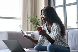 Serious young black African woman holding paper document calculating rent or money savings, paying bills in mobile application on cell phone doing monthly paperwork sitting on couch at home.
