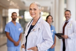 Portrait of mature female doctor on hospital corridor. Confident general practitioner standing in hospital hallway with her healthcare team in background. Successful head physician in private clinic.