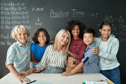 Portrait of cheerful smiling teacher and diverse schoolchildren standing posing on chalkboard blackboard background looking at camera happy after school reopen on mathematics lesson. Diversity.
