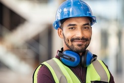 Portrait of satisfied construction site manager wearing safety vest and blue helmet with copy space. Young middle eastern architect watching construction site with confidence and looking at camera.