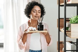 home improvement, decoration and people concept - happy smiling woman smelling aroma reed diffuser on board with cotton flower at shelf