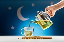 bedtime and sleeping concept - close up of hand pouring chamomile tea from teapot into mug at night over moon and night stars on blue background
