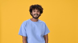 Smiling young curly indian cool positive guy standing isolated on yellow background. Happy ethnic stylish millennial man wearing blue casual t-shirt looking at camera posing for portrait.