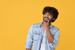 Smiling curious young indian man holding hand on chin looking interested aside at copy space isolated on yellow background thinking of shopping opportunities, planning purchase or dreaming concept.