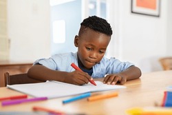 Little black boy sitting on table and painting with colored marker on book. Cute african american child drawing in the living room at home. Schoolboy colouring in book with red marker at home.