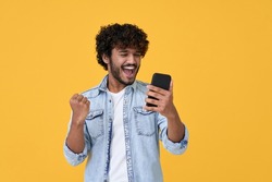Excited happy young indian man winner feeling joy using smartphone winning lottery game, betting, getting cashback online gift in mobile app message holding cell phone isolated on yellow background.