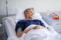Elderly patient sleeping on a medical bed in hospital ward. Senior woman resting after operation with eyes closed and IV drip. Old mature woman lying on bed and wearing blue hospital gown.