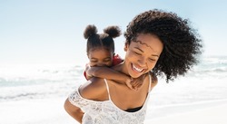 Happy black mother giving piggyback ride to her daughter at beach while laughing. Close up of beautiful and carefree mom carrying on shoulders her kid. Cute african little girl with bigger sister play