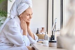 Attractive mid age older adult 50 years old blonde woman wears bathrobe and towel in bathroom touching face, looking at mirror doing daily beauty routine. Skin care treatment concept.