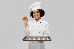 cooking, culinary and bakery concept - happy smiling female chef or baker in white toque and jacket holding baking tray with oatmeal cookies over grey background