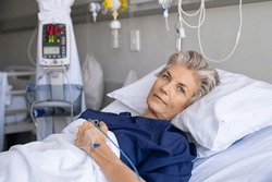 Mature woman recovering from illness while lying on hospital bed with iv drip in hand. Portrait of old hospitalized patient recovering after surgery. Happy senior woman lying on bed in hospital ward.