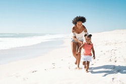 Happy black mother with cute little girl running barefoot on white tropical beach. Cheerful african american woman running along beach with her cute little sister. Smiling mother and funny female kid.
