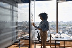 Young African american executive manager businesswoman writing strategy ideas on sticky notes on whiteboard doing business project presentation in contemporary corporate office with panoramic view.