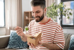 consumption, delivery and people concept - smiling man with fork and knife eating takeaway food at home