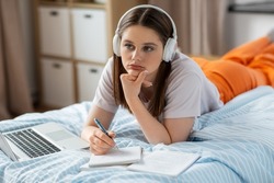 school, online education and e-learning concept - thinking teenage student girl in headphones with laptop computer writing to notebook lying on bed at home