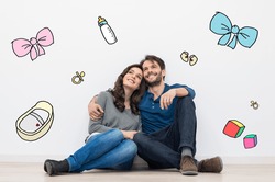 Portrait of happy young couple sitting against a white wall and dreaming to have a baby and a family. Their dreams are sketched with colors on the wall.