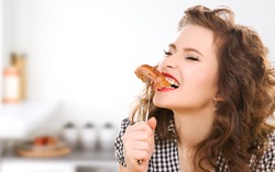 people, diet, culinary and food concept - hungry young woman eating meat on fork over kitchen background