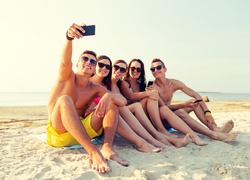 friendship, leisure, summer, technology and people concept - friends sitting and taking selfie with smartphone on beach