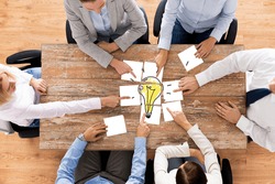 business, office people, startup and teamwork concept - close up of creative team sitting at table and putting together puzzle pieces with light bulb picture