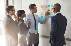 Multiracial group of colleagues discussing a business plan standing around a set of colorful memo notes stuck on the wall