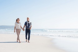 Senior couple holding hands at the beach on a sunny day. Mature couple in love holding hands and looking each other at the seaside. Smiling wife and happy husband walking barefoot on the white sand.