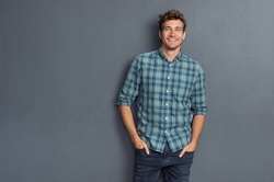 Handsome young man on grey background looking at camera. Portrait of laughing young man with hands in pockets leaning against grey wall. Happy guy smiling.