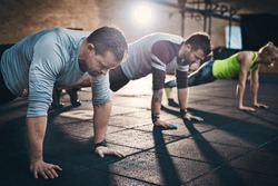 Group of adults performing push up exercise drills at indoor physical fitness cross-training exercise facility with bright light flare over them