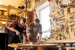 profession, carpentry, woodwork and people concept - two carpenters with electric drill drilling wood plank at workshop