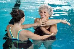 Young trainer helping senior woman in aqua aerobics. Senior retired woman staying fit by aqua aerobics in swimming pool. Happy old woman stretching in swimming pool with young trainer.