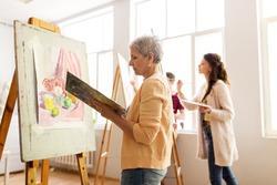 art school, creativity and people concept - happy senior woman artist with easel, paint brush and palette painting at studio