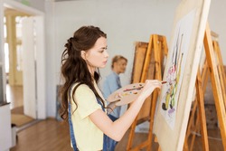 art school, creativity and people concept - student girl or young woman artist with easel, palette and paint brush painting still life picture at studio