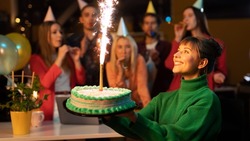 Beautiful Caucasian Female Holding a Cake as Her Co-Workers Cheer Her on In The Background Wearing Party Hats. Caucasian Office Workers Throwing a Surprise Birthday Party For Their Co-Worker.