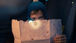 Close Up Shot of a Caucasian Young Boy Holding the Treasure Map and a Flashlight, Looking Serious As He Tries to Decipher the Code, Sitting and Hiding in a Dark Tent During The Night.