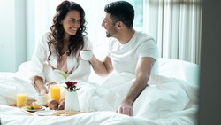 Isolated Beautiful Ethnic Couple Celebrating Valentines Day in a Fancy Hotel Suite, Eating Breakfast Off of a Fancy Tray As They Stay Under The Blanket, Looking Into Each Others Eyes and Smiling.