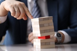 Close-up view of company leader pressing on wooden tower on one hand. Skewed pyramid of light blocks with one red brick in the middle. Risk and vulnerable situation concept