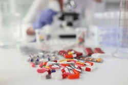 Pile of pills scattered on white table against woman doing research with microscope. Medicine and scientific research in laboratory