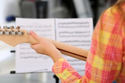 Female hand holding wooden neck of electric guitar and playing song using note papers in music stand closeup. Learning musical instrument, music shop or school, having fun enjoying hobby concept