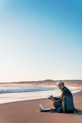 digital nomad sitting on the beach with his laptop and taking notes diary on the shore of the beach at sunset