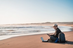 digital nomad sitting on the beach working with his laptop on the shore of the beach at sunset