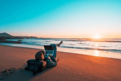 mature latin digital nomad laying on the sand of the beach with a laptop working at sunset over the ocean