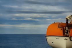 Lifeboat hanging above the decks onboard a cruise ship. Orange and white raft for use in a nautical emergency. With view of the sea with a moody sky. Space for text.